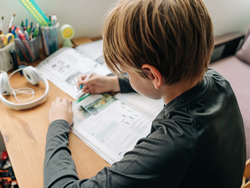 Kind sitzt vor seinem Pult, auf welchem Kopfhörer, Stifte und Arbeitsbücher liegen.
