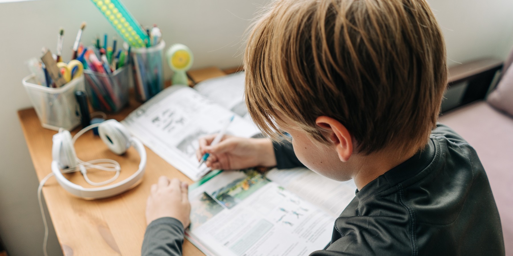 Kind sitzt an seinem Pult, auf welchem Stifte, Arbeitshefte und ein Paar Kopfhörer liegen.